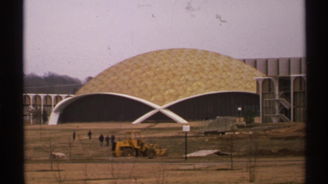 Gold geodesic dome building with white arched front, construction dirt, small pedestrians, and a yellow loader.