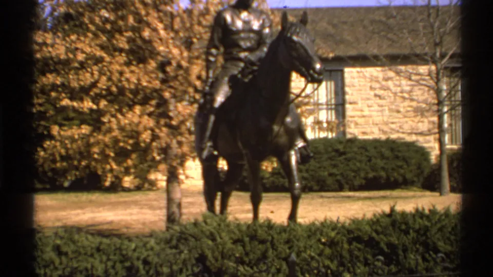 1972 Tulsa 8mm frame of a bronze equestrian statue in landscaped grounds, with autumn foliage, hedges, lawn, and a stone wall behind it.