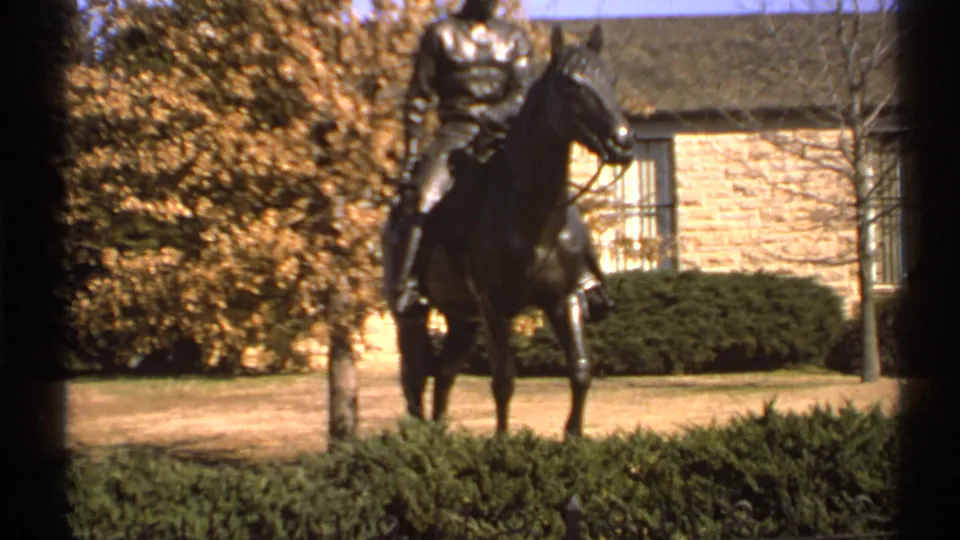 1972 Tulsa home-movie frame showing an equestrian statue before manicured hedges, autumn trees, lawn, and a stone building facade.
