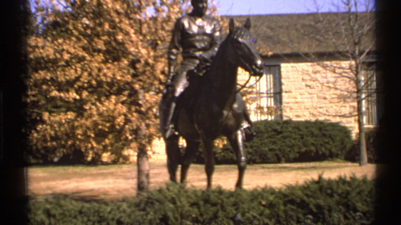 Bronze equestrian statue near autumn trees and a low stone building.