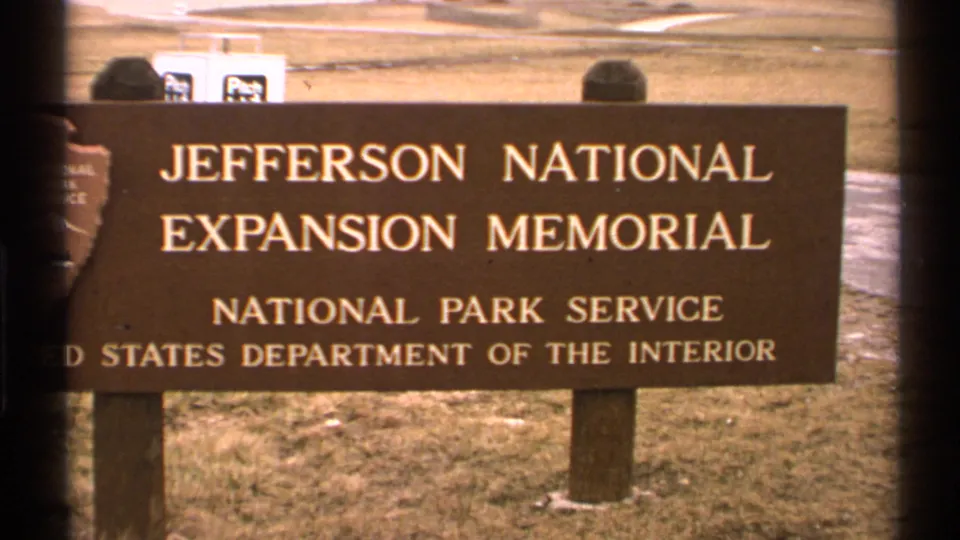 A brown sign reads Jefferson National Expansion Memorial and National Park Service, with grass, sidewalk, and a partial visitor visible behind it.