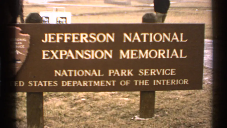 The brown Jefferson National Expansion Memorial sign remains centered, with official text, grass, and a partial passerby behind the sign.