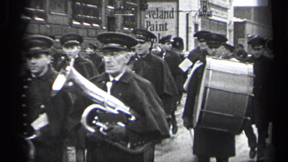 A Cleveland marching band advances along the street, with a trombone player in front and a bass drummer visible near a Cleveland Paint sign.