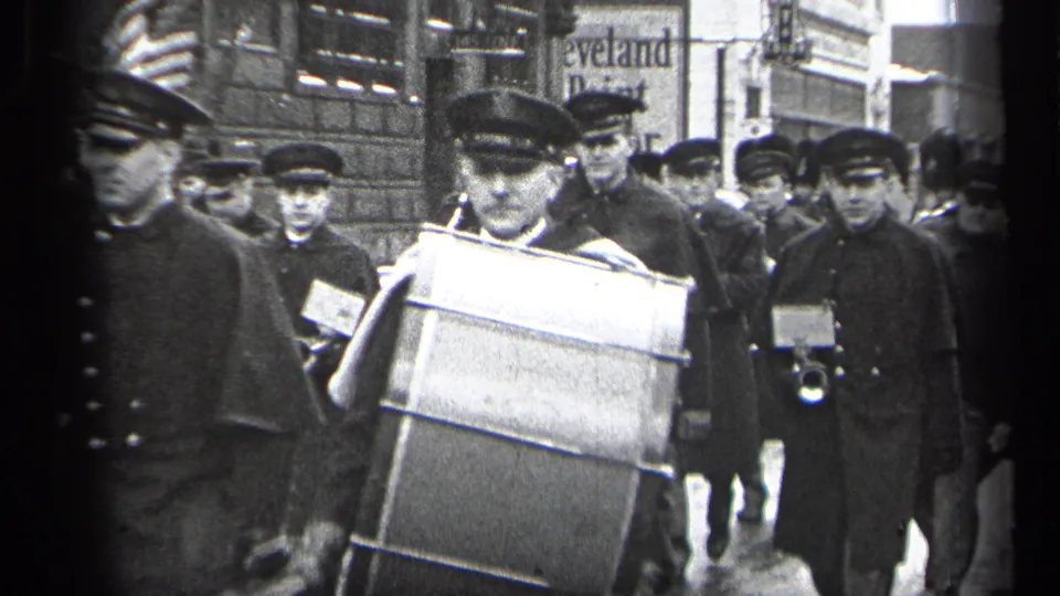Close view of a uniformed bass drummer in the 1937 Cleveland procession, with additional musicians and street spectators behind him.