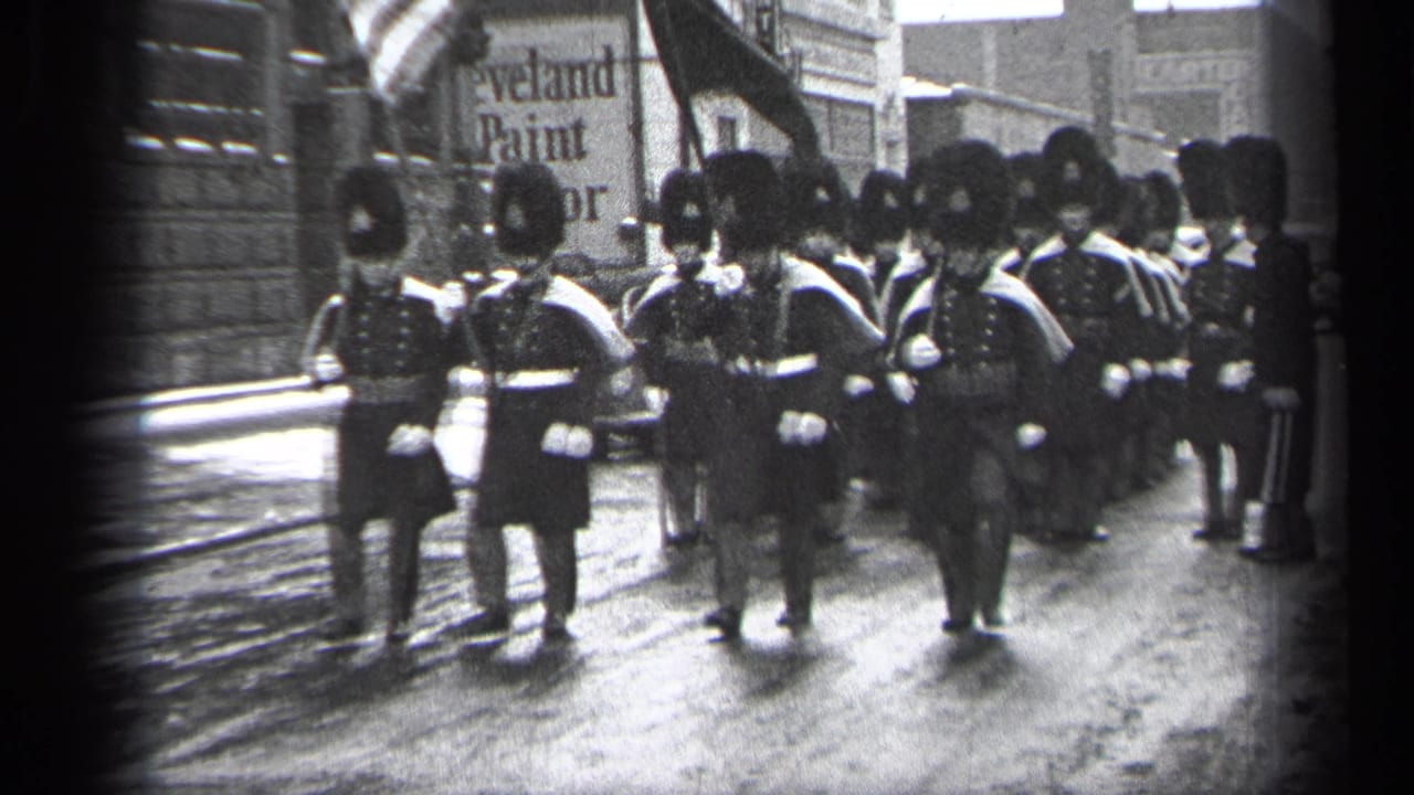Uniformed marchers in tall hats advance along a wet city street past storefronts.