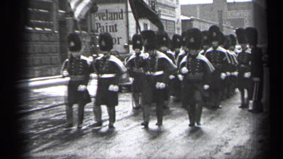 In 1937 Cleveland, uniformed marchers in tall dark hats and white gloves move toward the camera on a wet commercial street.
