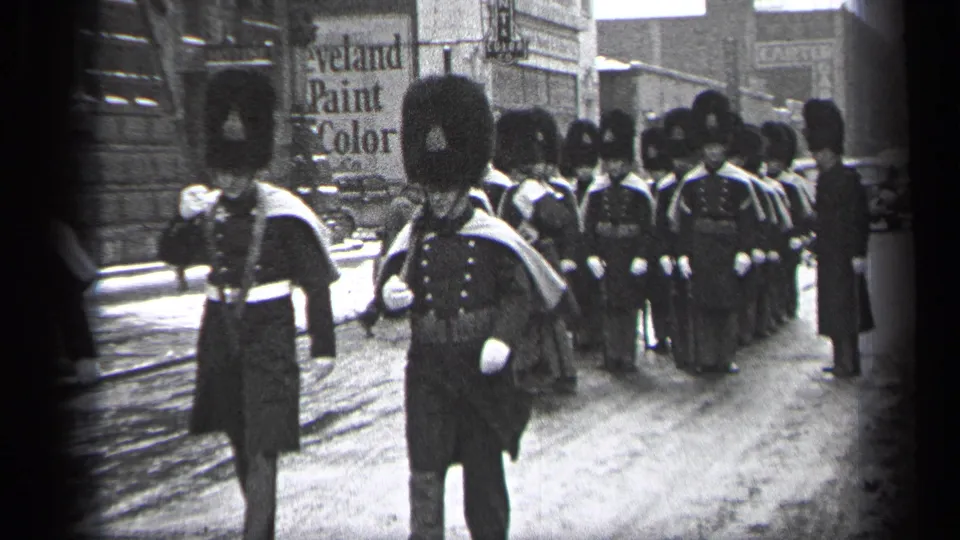 A close street-level view shows two uniformed marchers passing the camera as the parade line gathers behind them in Cleveland.