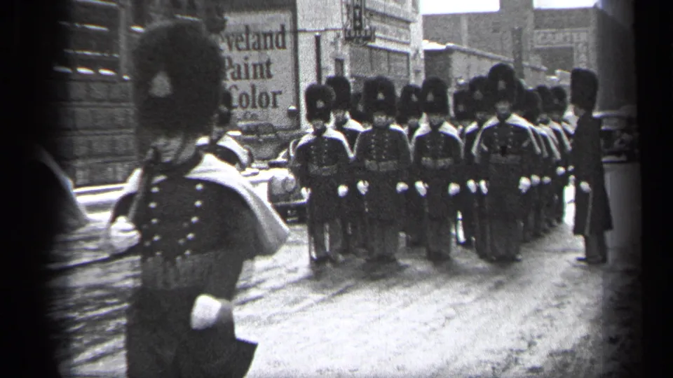 A foreground marcher moves across the frame as uniformed parade participants stand in formation on the wet Cleveland street.