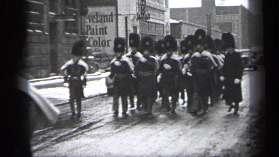1937 Cleveland home-movie frame showing a uniformed parade group in tall dark hats marching past storefronts and parked cars.