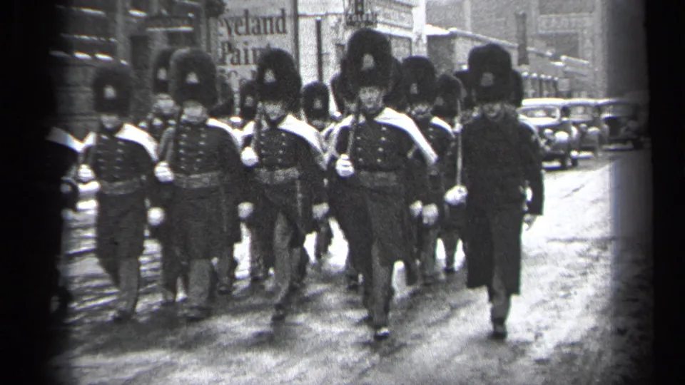 Close 1937 Cleveland frame of uniformed parade marchers with tall hats, white gloves, and a suited man at the side.