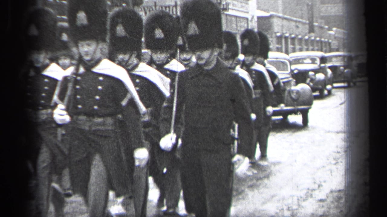 Uniformed ceremonial marchers pass along a city street beside 1930s cars.