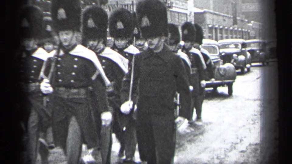 In Cleveland according to metadata, uniformed marchers in tall ceremonial hats move past storefronts and parked or moving 1930s automobiles.