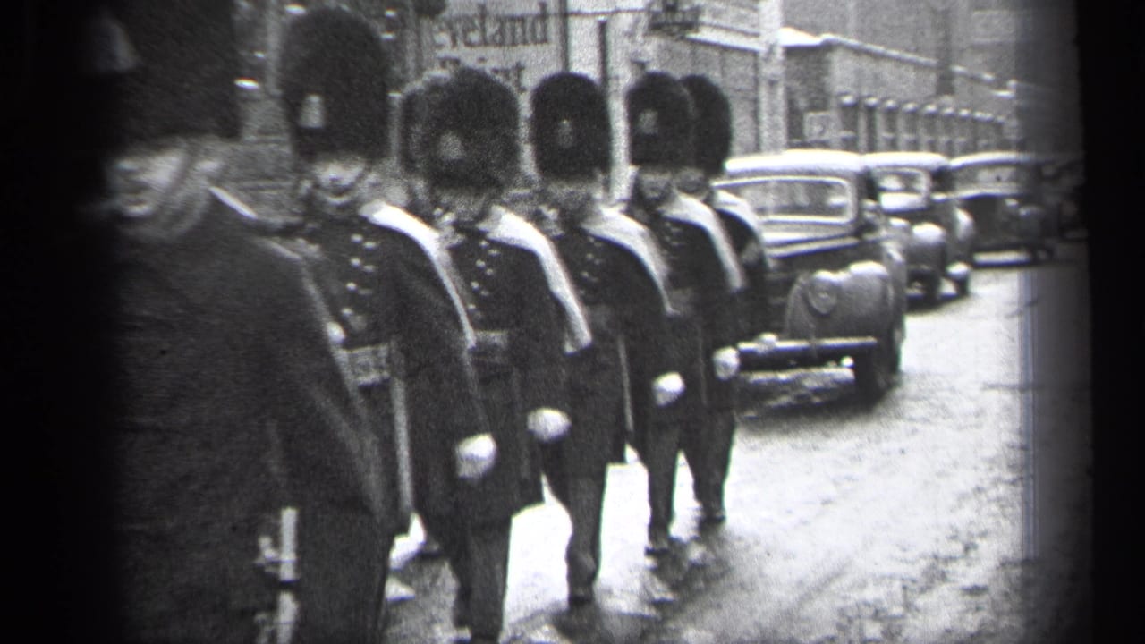 Marching soldiers in tall hats pass close to the camera as cars move behind them.