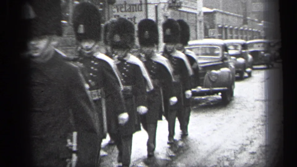 Ceremonial soldiers in dark uniforms and tall hats pass near the lens on a Cleveland street, with classic sedans visible in the roadway.