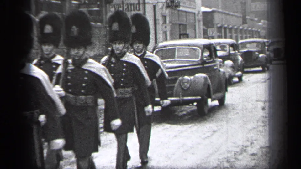 A curbside 8mm view shows ceremonial marchers crossing the frame in Cleveland, with 1930s cars and storefront signs behind them.