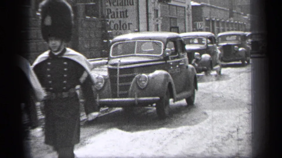Near the end of the clip, a uniformed marcher stands partly at frame edge while a prominent 1930s sedan passes along the Cleveland street.