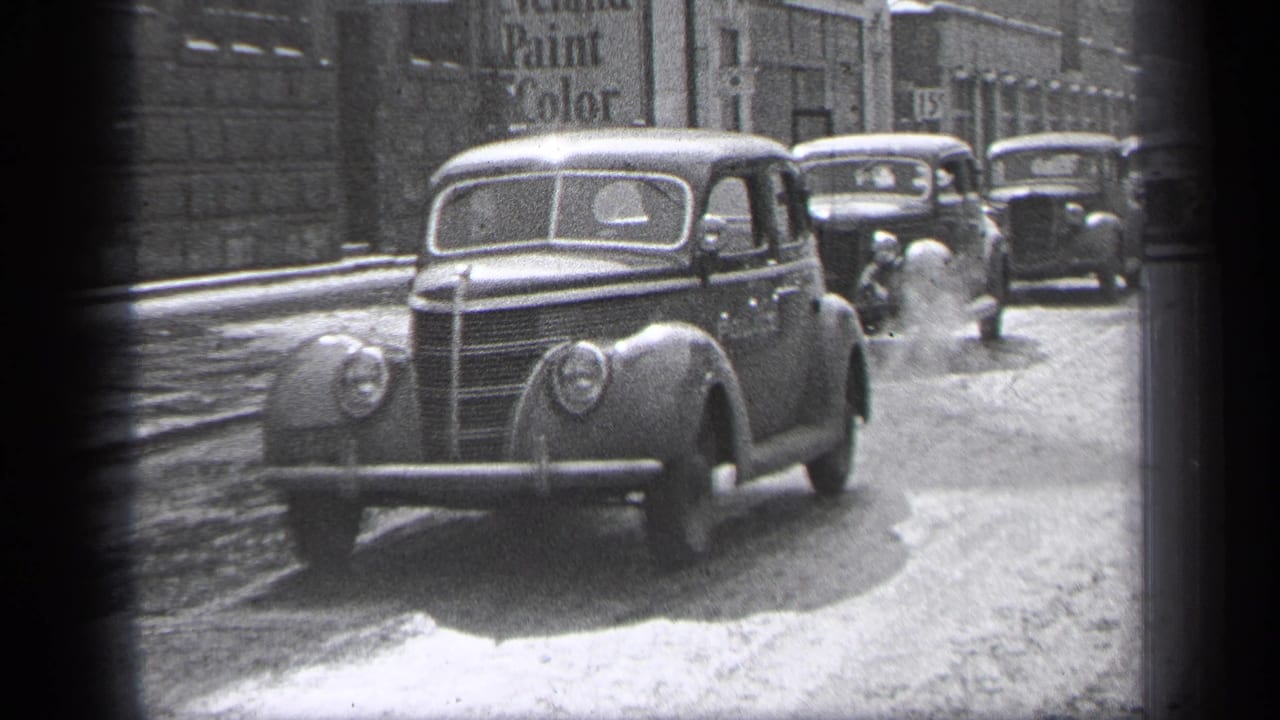 Classic 1930s cars on a city street with buildings and a painted sign in the background.