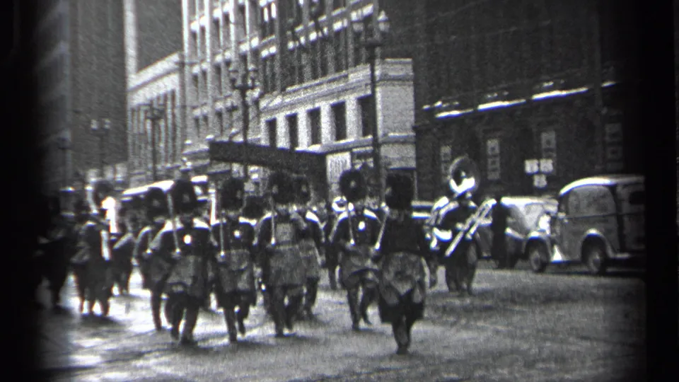 Marching band in formal uniforms moves through a 1937 Cleveland street with parked or moving automobiles and urban buildings behind.