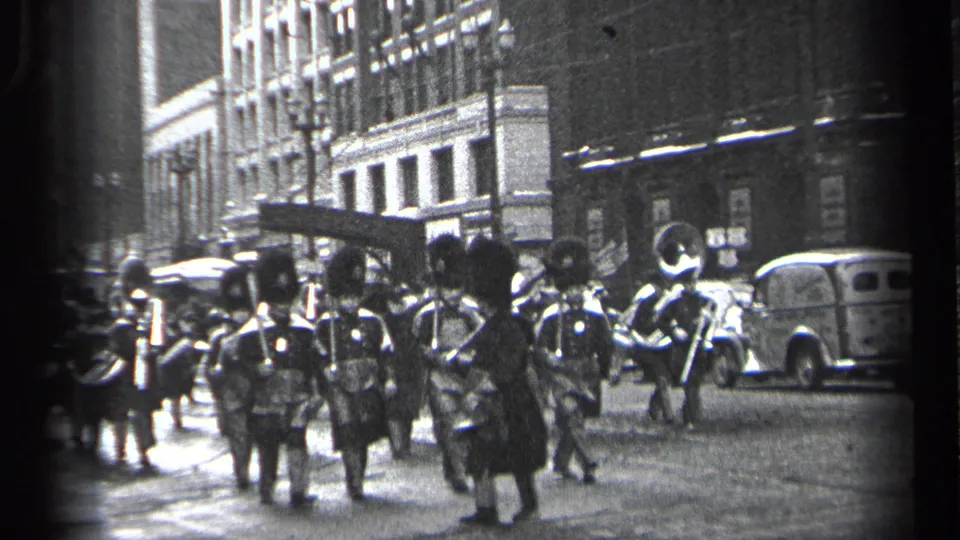 Cleveland parade view with marching band members in tall hats moving past automobiles and spectators on a downtown street.