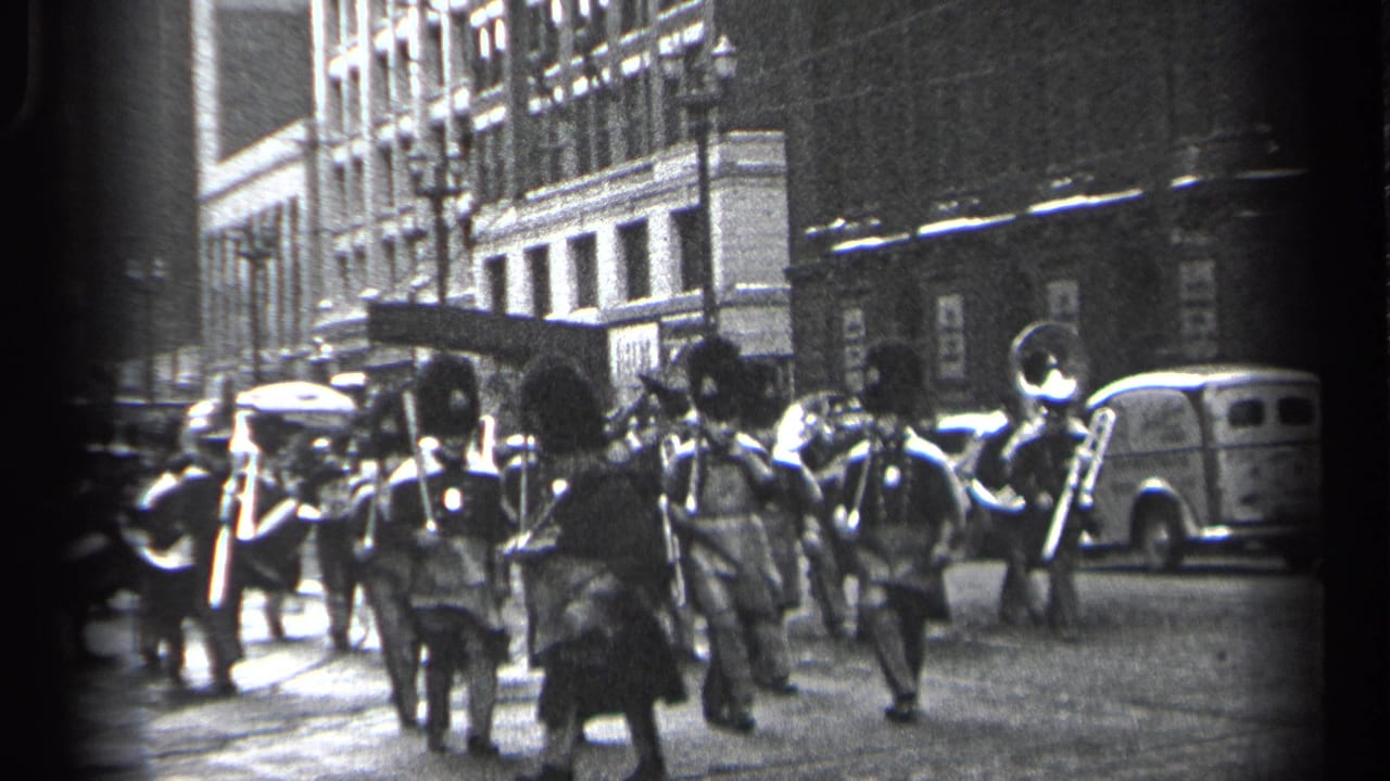 In Cleveland, Ohio, 1937, a uniformed marching band advances along a city street as pedestrians and parked cars line the curb.