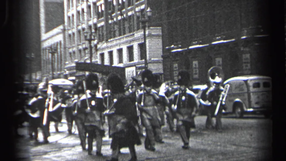In Cleveland, Ohio, 1937, a uniformed marching band advances along a city street as pedestrians and parked cars line the curb.