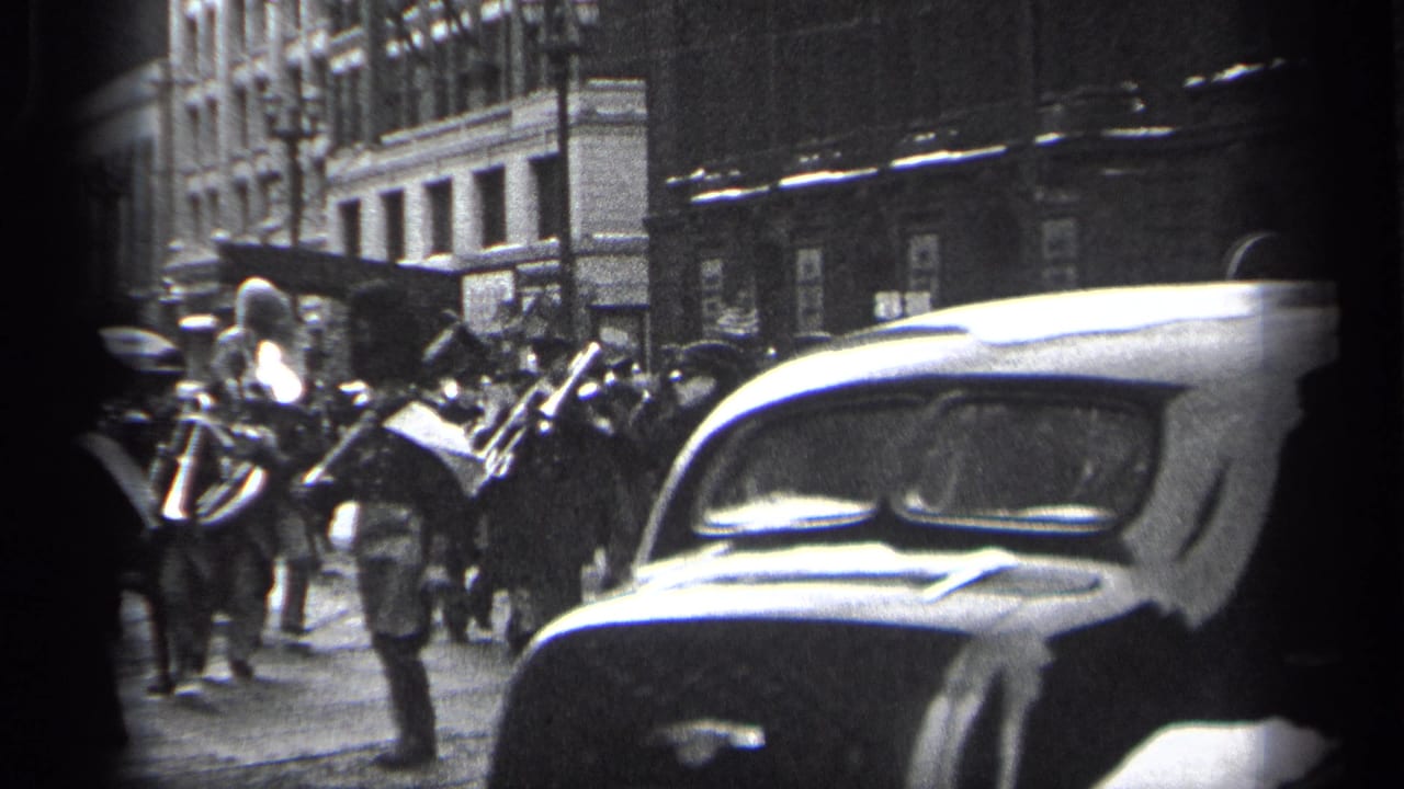 Marching band passes on a city street as a large 1930s car crosses the foreground.