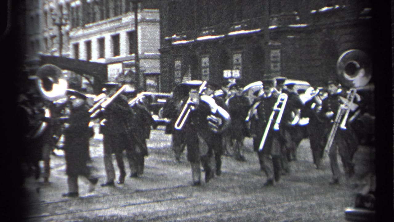 Uniformed band members march with tubas and trombones along a city street.