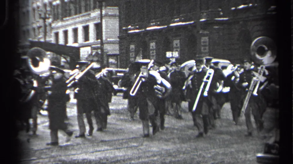 Cleveland 1937 street parade frame showing brass players in dark uniforms moving past parked cars and urban buildings.