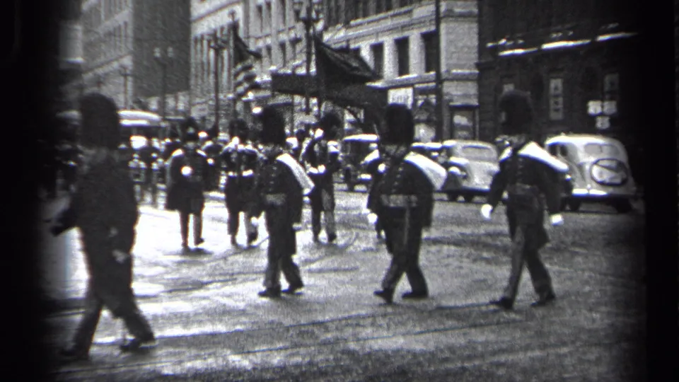 Cleveland 1937 parade frame showing honor guard marchers, flags, pedestrians, parked cars, and city buildings.