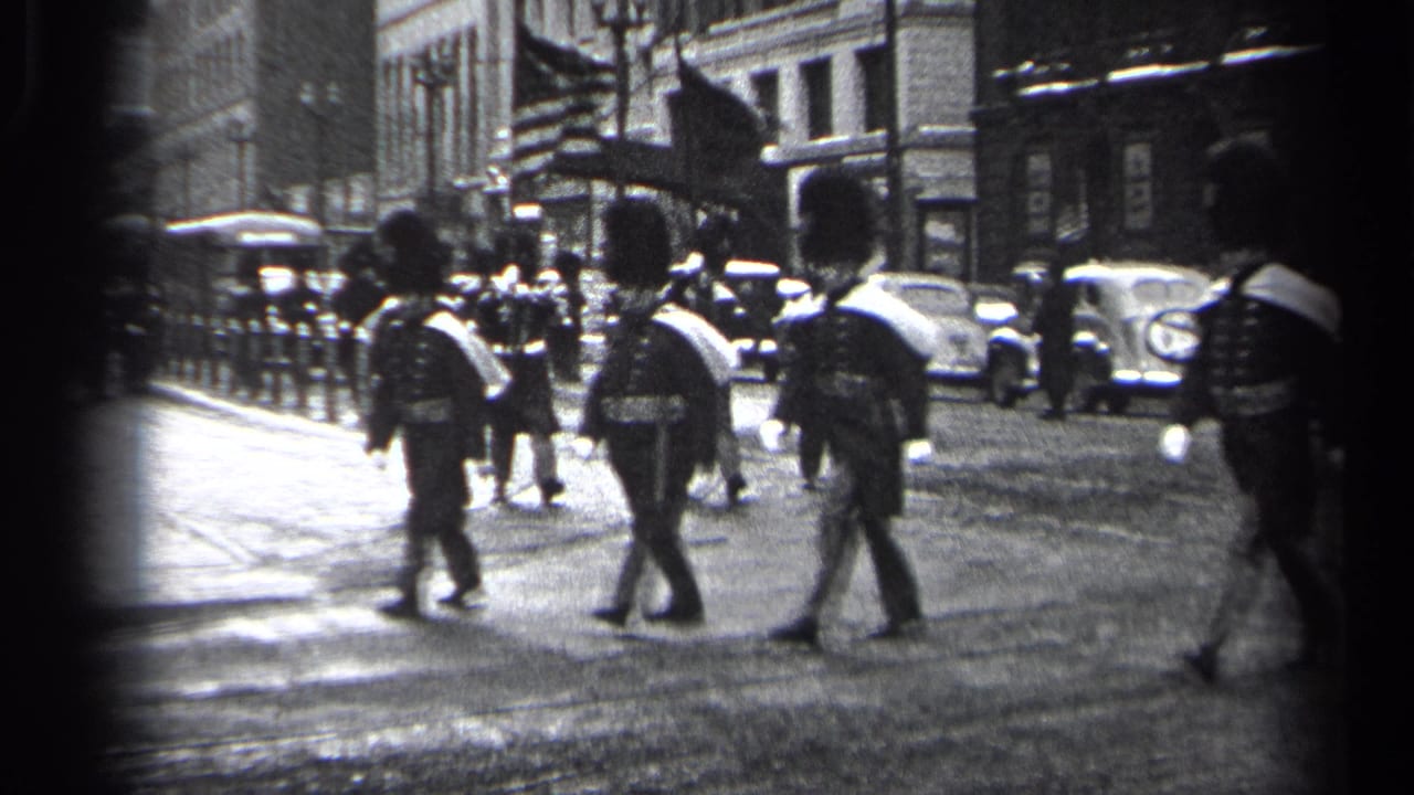 Uniformed marchers in tall hats cross a city street with parked cars and buildings behind them.