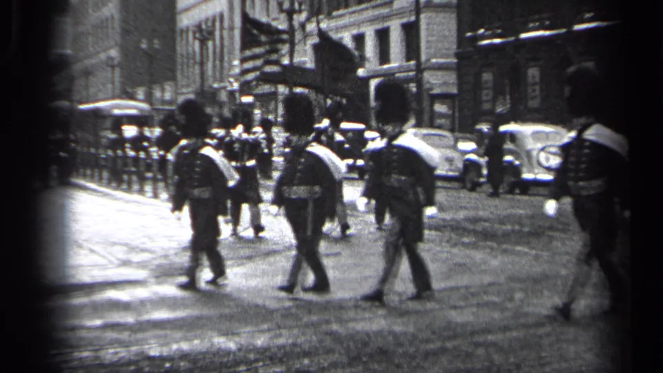 In 1937 Cleveland, uniformed marchers with tall hats and white sashes move across an urban street lined with parked automobiles.