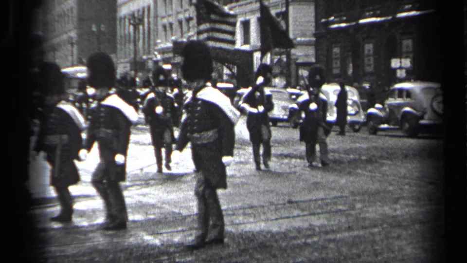 Uniformed procession members continue down a Cleveland street in 1937, with older cars, spectators, and building fronts in the background.