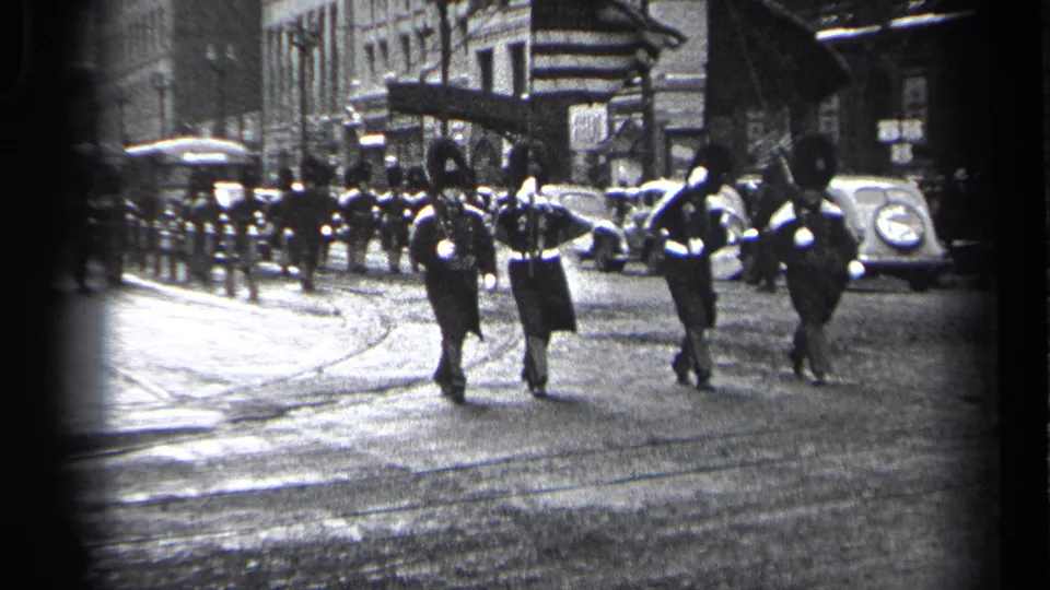 A small group of uniformed marchers moves away from the camera in 1937 Cleveland, with flags, parked cars, and curbside viewers visible.