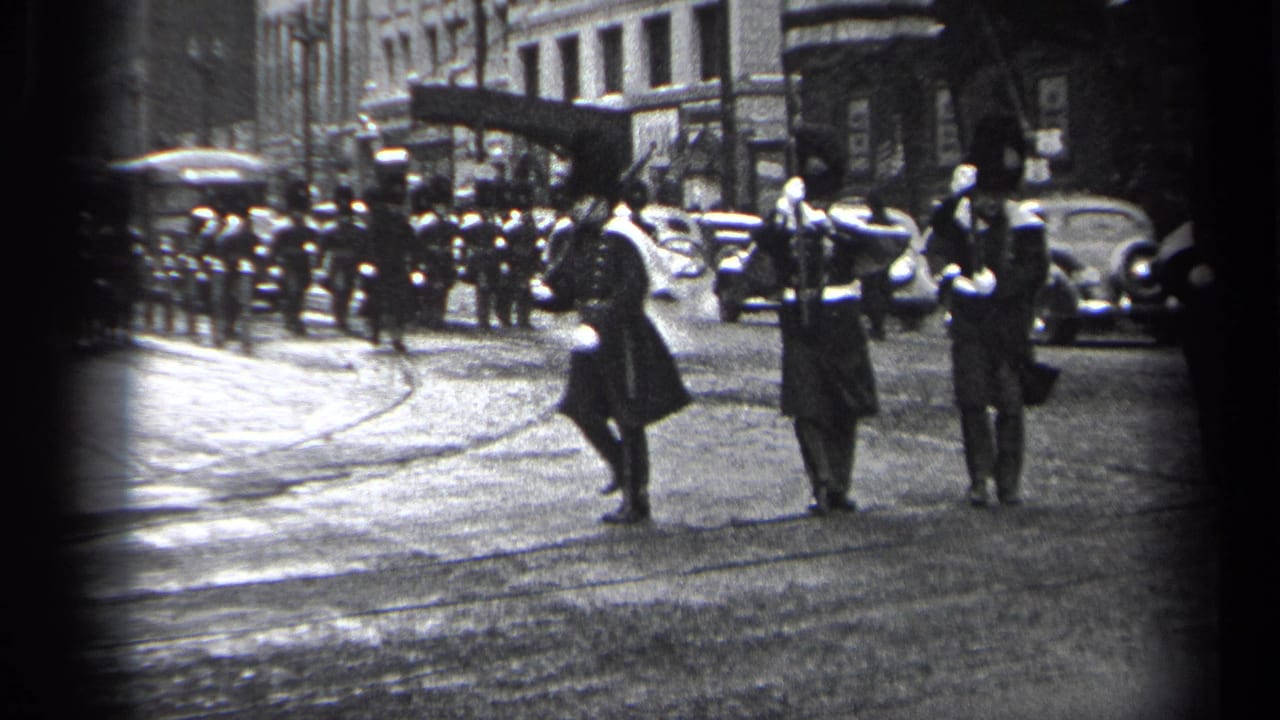 In 1937 Cleveland, uniformed men in tall hats march across a city street with parked cars, spectators, and masonry buildings behind them.