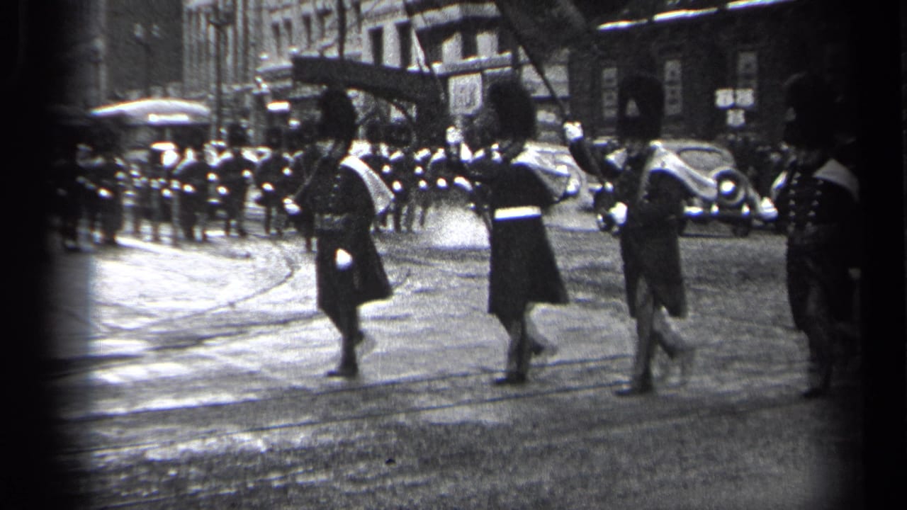 Three uniformed men march past the camera with spectators and cars in the background.