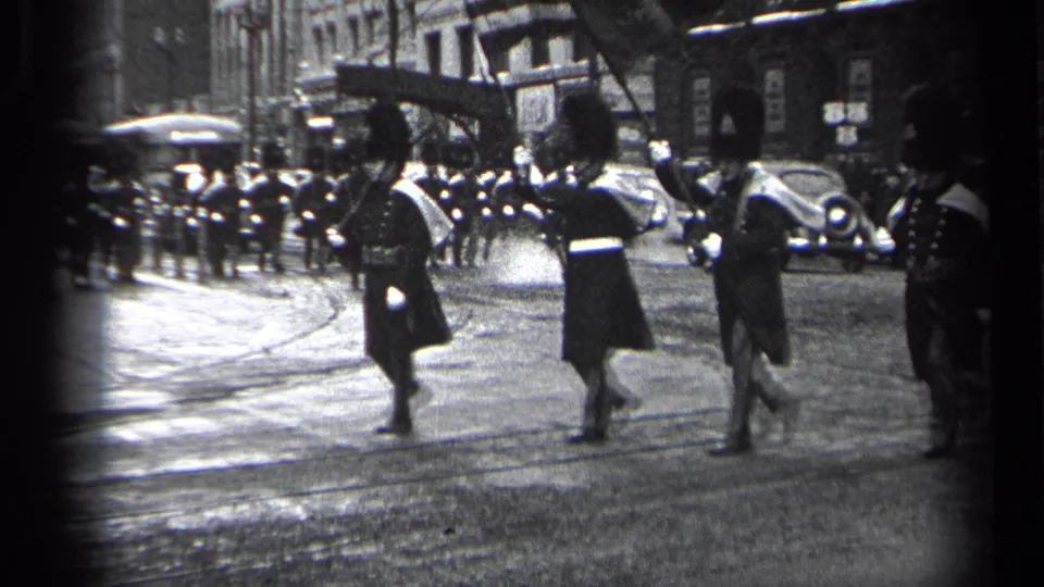 Uniformed marchers with tall hats and white cross-belts pass through a Cleveland street scene with 1930s cars and onlookers visible behind them.
