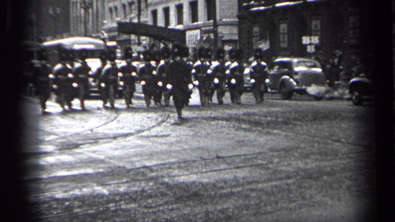 Uniformed marchers cross a wet city street with cars and buildings behind them.