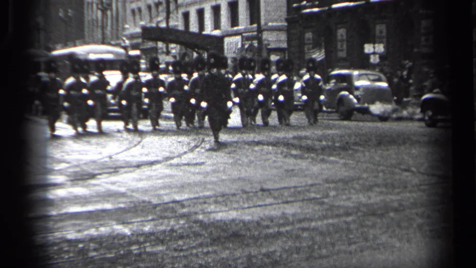 1937 Cleveland home-movie frame showing uniformed marchers moving across a wet street with period cars and buildings in the background.