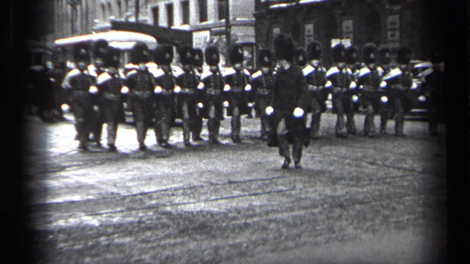 1937 Cleveland 8mm frame showing uniformed marchers in tall hats forming across an urban street with buildings and vehicles behind them.