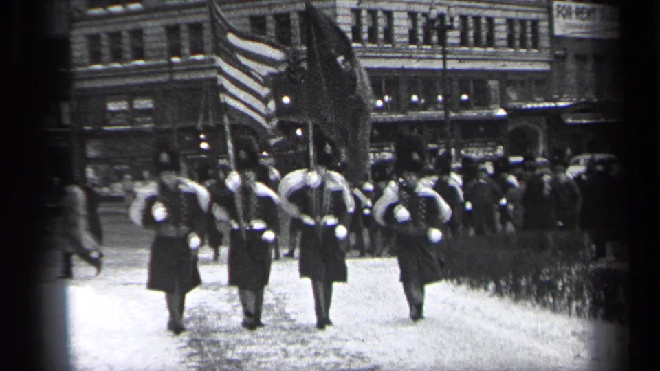 Uniformed flag bearers in 1937 Cleveland march away from the camera, with commercial buildings, spectators, and street traffic nearby.