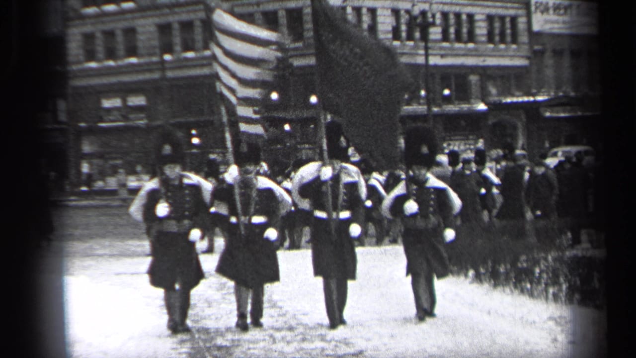 In Cleveland, Ohio, 1937, uniformed men in tall hats march with flags and rifles past downtown buildings.