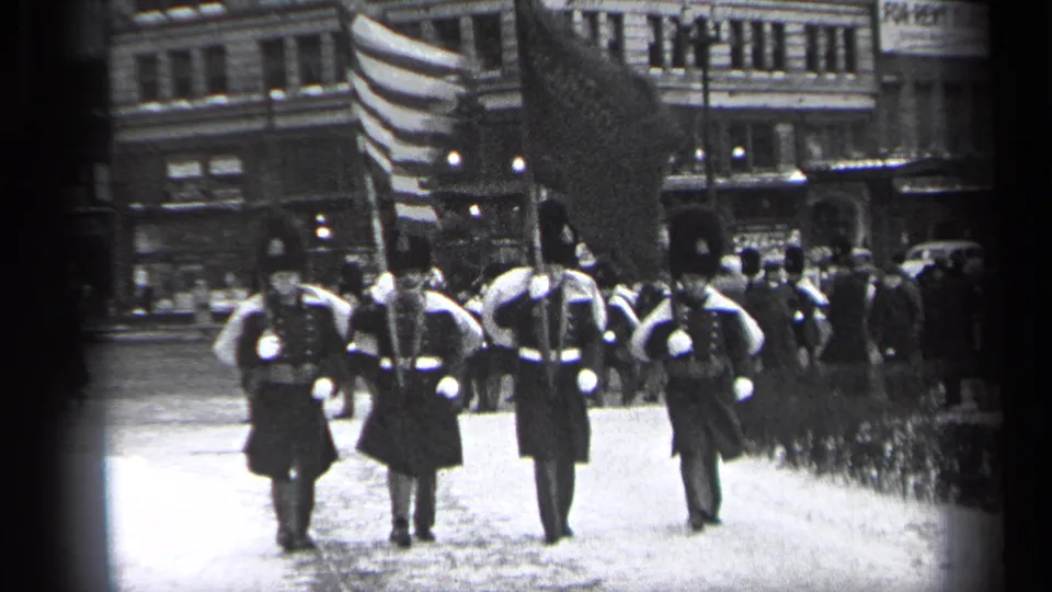 In Cleveland, Ohio, 1937, uniformed men in tall hats march with flags and rifles past downtown buildings.