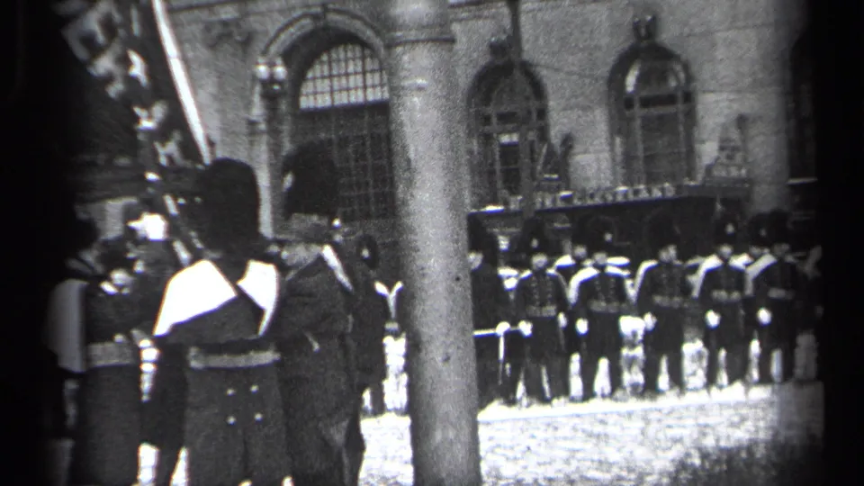 A soldier in formal dress stands near a flag or standard as rows of uniformed personnel line up outside the arched building.