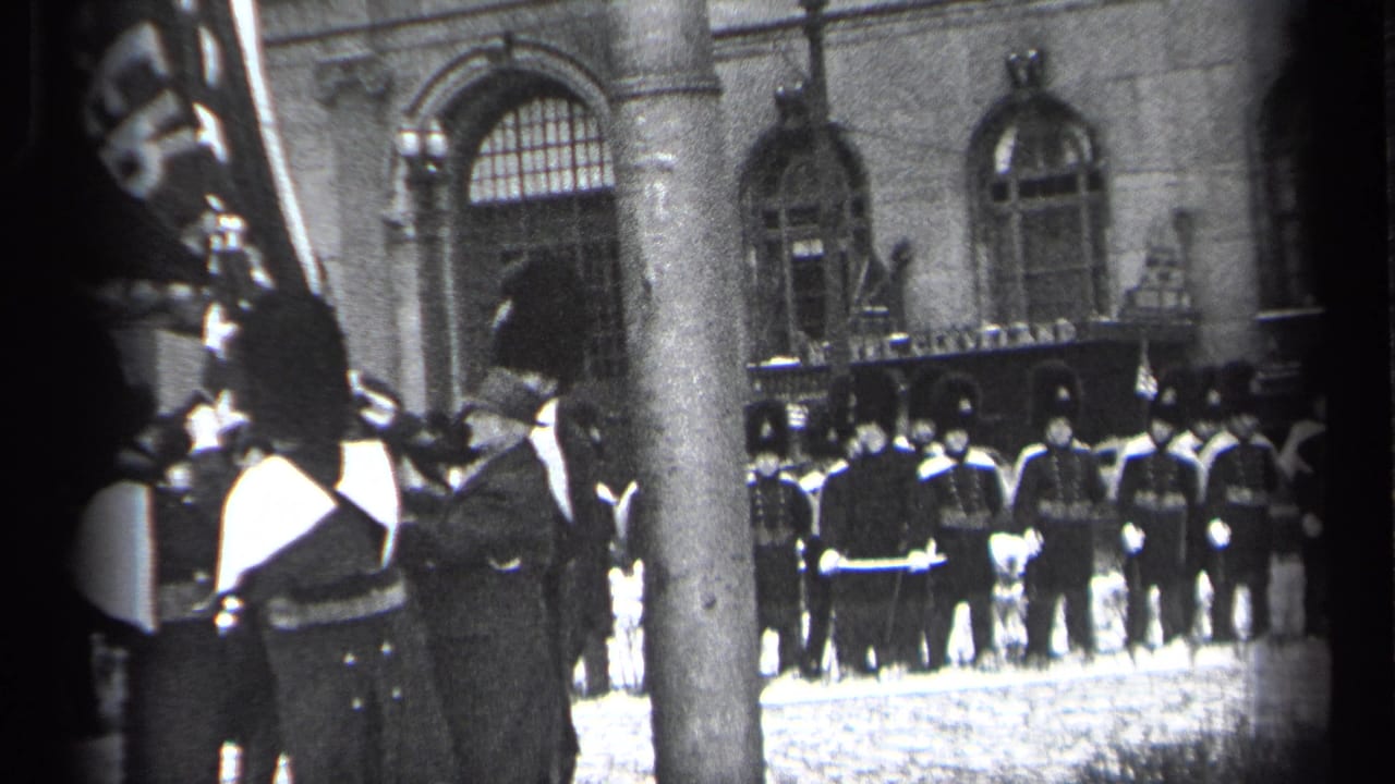 Uniformed soldiers stand in formation outside a stone building while spectators watch from the foreground.