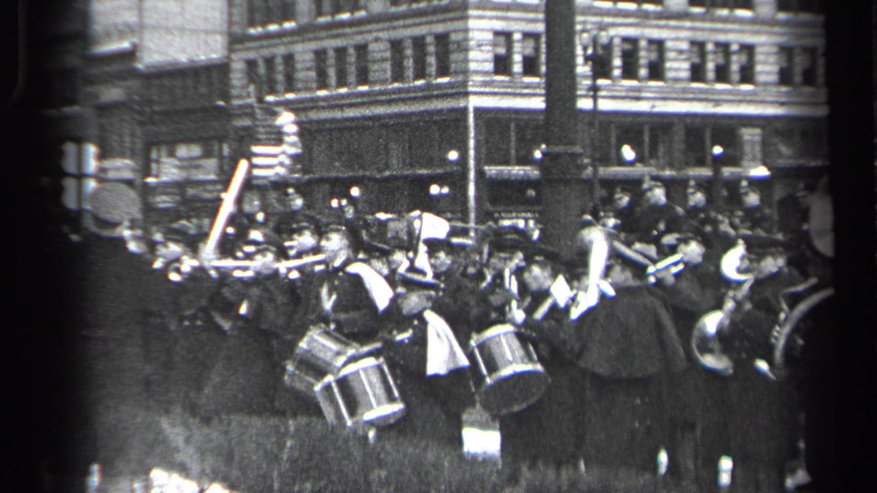 Uniformed marching band members play drums and brass in a crowded city street parade.