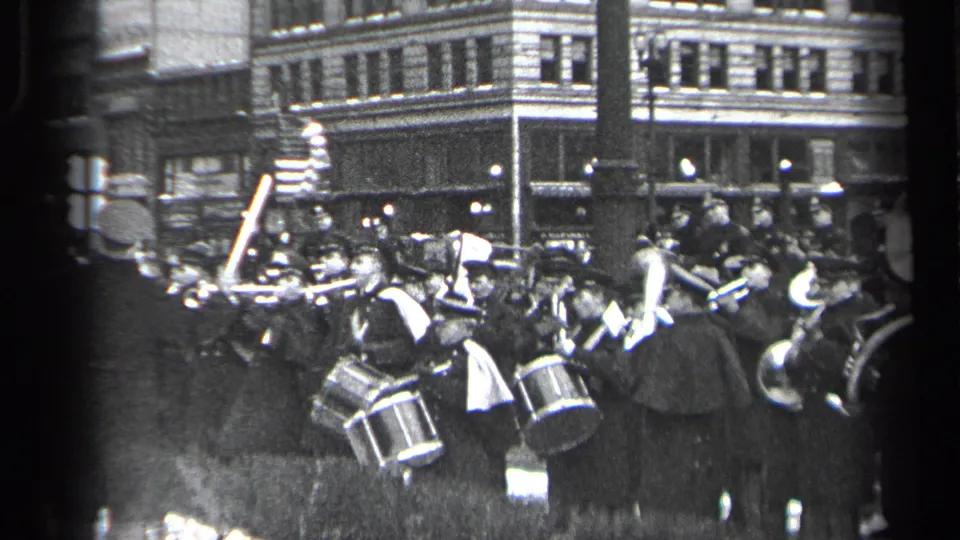 1937 Cleveland street parade view with uniformed drummers, brass players, spectators, an American flag, and commercial buildings behind them.