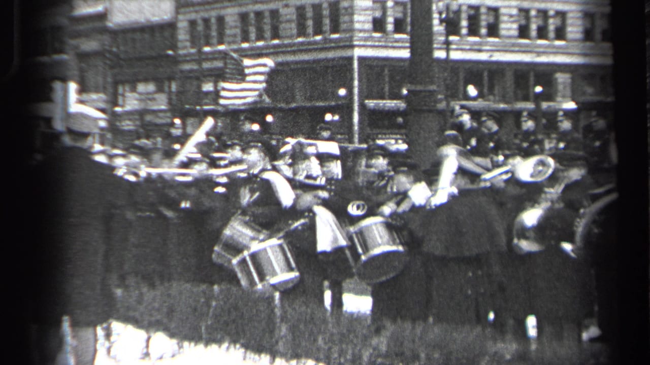 Band members with drums and horns move through a parade crowd on a city street.