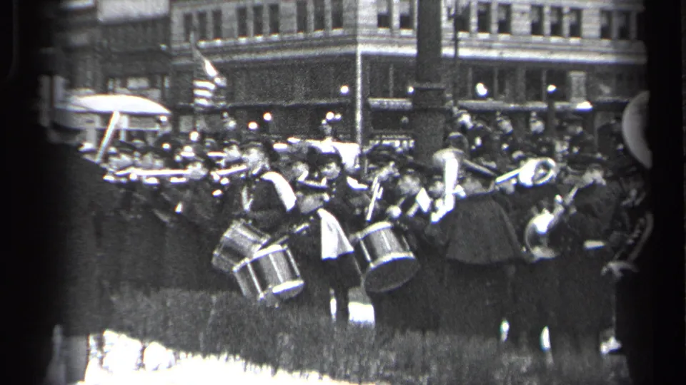 Black-and-white 8mm frame of a Cleveland marching band in 1937, with drums and horns visible amid a crowded street scene.
