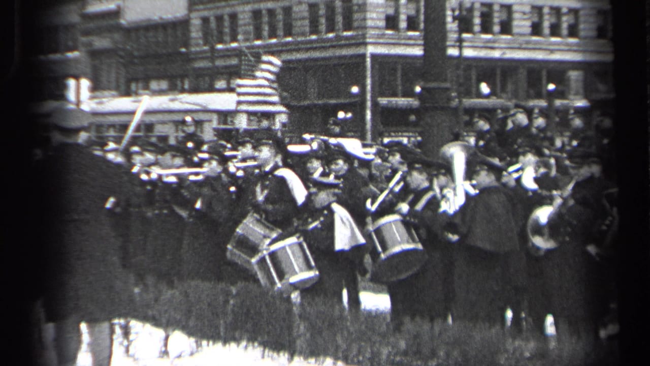 Marching band plays in a city street with drums and brass instruments near spectators.