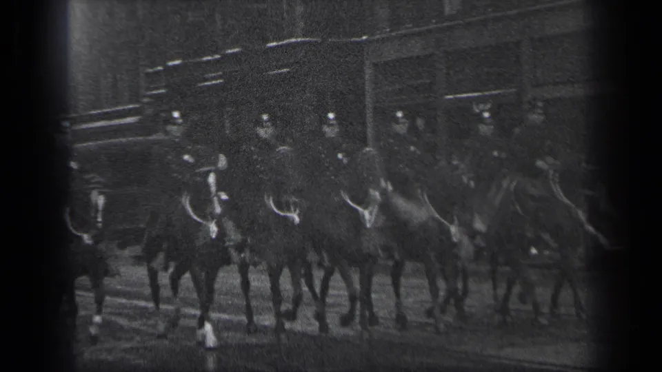 In 1937 Cleveland, mounted police officers in dark uniforms ride horses past urban buildings in grainy 8mm footage.